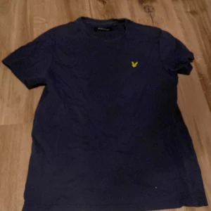 Mörkblå t-shirt från Lyle & Scott - Säljer en mörkblå t-shirt från Lyle & Scott med klassisk gul logga broderad på bröstet. T-shirten har rund hals och korta ärmar. Perfekt för en avslappnad stil.