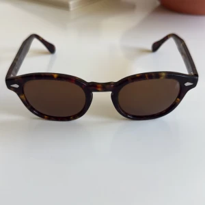 Bruna solglasögon från Moscot Originals - Snygga solglasögon från Moscot Originals i klassisk rund form. Bågarna har ett brunt sköldpaddsmönster och glasen är mörktonade. Perfekta för soliga dagar och ger en tidlös look.