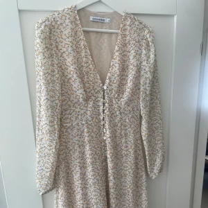 Adoore klänning Paris lemon - Säljer en vacker långklänning från Adoore med små blommor i gult och beige på vit botten. Klänningen har v-ringning, lång ärm och knappar framtill. Perfekt för sommarens tillfällen och har en feminin passform. Sparsamt använd och i fint skick utan defekter.