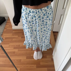 Vit blommig kjol med volangkant - Supersöt vit kjol med blått blommönster och volang längst ner. Kjolen har resår i midjan och är i en längre modell som känns både romantisk och somrig. Perfekt att styla med en enkel topp eller stickad tröja.🥰