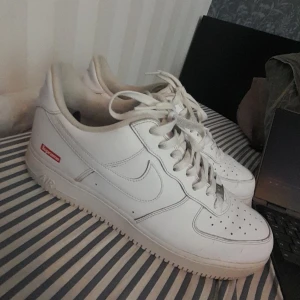 Nike Air Force 1 x Supreme vita sneakers - Säljer ett par vita Nike Air Force 1 Supreme-logga samarbete med Supreme. Skorna har klassisk låg siluett, vit läderöverdel, Supreme-logga på sidan och Nike Swoosh.