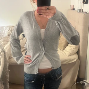 Grå trendig kofta - Säljer en klassisk grå kofta från KappAhl med v-ringning och knäppning framtill. Koftan har långa ärmar och ribbade muddar vid ärmslut och nederkant. Perfekt att slänga över en t-shirt/linne tillsammans med ett par jeans!