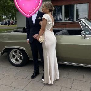 Champagnefärgad långklänning med öppen rygg - Elegant långklänning i champagnefärg med djup v-ringning och öppen rygg med knyt. Klänningen har korta ärmar och är figurnära med släp. Perfekt för bal eller festliga tillfällen.