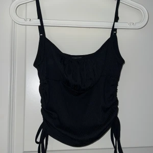 Svart ribbad linnetopp med knyt - Säljer en svart ribbad linnetopp med smala axelband och knyt i sidorna. Toppen har en lätt rynkad detalj framtill och är croppad i modellen. Perfekt att styla till jeans eller kjol. Storlek xs ( från Shein )