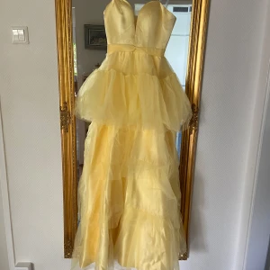 Gul volangklänning med sweetheart-urringning - Superfin balklänning i ljusgul färg med flera lager volanger och en snygg urringning. Axelbandslös modell som ger en elegant och drömmig känsla, perfekt för bal eller fest. Klänningen har markerad midja och är tillverkad i ett lätt, fluffigt material. Klänningen är aldrig använd o ja säljer den för den tyvärr inte satt så bra på mig! ❣️