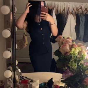 Marinblå set med knappar - Säljer ett marinblå set i tvådelad look med ärmlös topp och matchande kjol. Toppen har dekorativa guldiga knappar framtill och rund halsringning. Materialet är mjukt och stretchigt, perfekt för en stilren och enkel look.