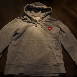 Grå hoodie från Comme des Garçons Play - Säljer en grå hoodie från Comme des Garçons Play med det ikoniska röda hjärtat med ögon broderat på bröstet. Tröjan har huva med dragsko och en stor magficka. Perfekt för en avslappnad och stilren look. Inte använd många gånger. Pris kan diskuteras!