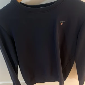 Mörkblå sweatshirt från GANT - En stilren mörkblå sweatshirt från GANT med rund halsringning och diskret logga på bröstet. Tröjan har långa ärmar och är perfekt för en avslappnad och klassisk look.