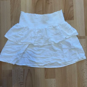 Vit volangkjol med resårmidja - Söt vit kjol med bred resår i midjan och dubbla volanger upptill. Kjolen är kort och har en luftig känsla, perfekt för varma dagar. Enkel att matcha med olika toppar för en fräsch look. Säljer pgå att den är för liten.