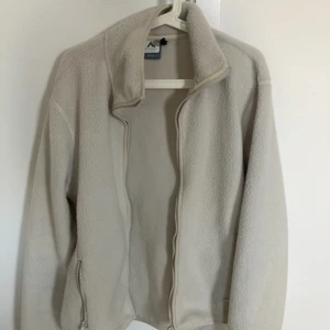 Beige fleecejacka med dragkedja - Mjuk och varm beige fleecejacka från Peak Performance med hög krage och hel dragkedja framtill. Jackan har två fickor med dragkedja och enkel, stilren design. Perfekt för kyliga dagar.