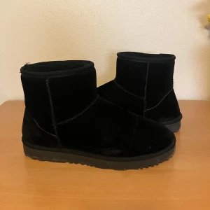 Svarta Uggs  - Säljer ett par klassiska svarta ”Uggs” från Nelly.  Skorna har en enkel och stilren design, perfekt för kyligare dagar. De är fodrade på insidan för extra värme och komfort.