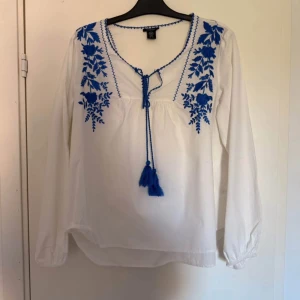 Vit broderad blus från Lindex  - Säljer en vacker, vit blus från Lindex i storlek M. Blusen har fina blå broderier och dekorativa detaljer vid halsen – perfekt för både vardag och festligare tillfällen. Den är helt oanvänd och i nyskick!