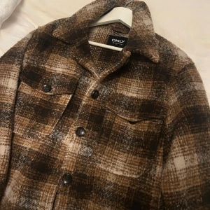 Brunrutig overshirt från ONLY - Snygg brunrutig overshirt från ONLY med stora knappar framtill och två bröstfickor. Jackan har klassisk krage och är perfekt att slänga över en hoodie eller t-shirt för en chill look.