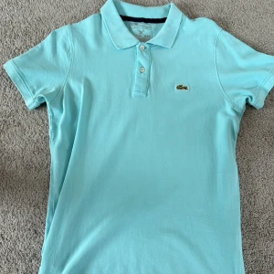 Ljusblå pikétröja från Lacoste - Säljer en klassisk ljusblå pikétröja från Lacoste med den ikoniska krokodilloggan broderad på bröstet. Tröjan har korta ärmar, krage och knappar framtill. Perfekt för en stilren och sportig look.