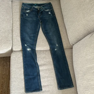 Levis Skinny jeans i mörkblå denim - Säljer ett par klassiska Levis Skinny jeans i mörkblå tvätt med slitningar och snygga detaljer på bakfickorna. Byxorna har normal passform och dragkedja framtill. Perfekta för dig som gillar en tidlös jeanslook.
