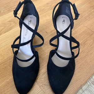 Svarta klackskor med remmar från Tamaris - Säljer ett par svarta klackskor från Tamaris med flera tunna remmar som korsar foten och spänne vid ankeln. Skorna har spetsig tå och är tillverkade i mocka. Köpta på Vinted men är oanvända av förra ägaren och mig. Skriv för fler bilder och priset går att diskuteras!