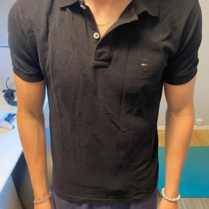 Svart pikétröja från Tommy Hilfiger - Säljer en klassisk svart pikétröja från Tommy Hilfiger med liten broderad logga på bröstet. Tröjan har korta ärmar, krage och knappar vid halsen. Perfekt för en stilren och avslappnad look.