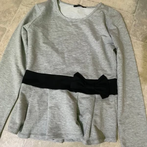 Ljusgrå långärmad topp med svart rosett - Säljer en ljusgrå långärmad topp med rund halsringning och en bred svart midjedetalj med rosett. Perfekt för dig som vill ha en enkel men ändå söt look. Toppens material känns mjukt och bekvämt.