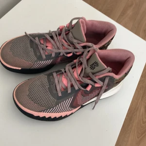 Nike Kyrie sneakers i grått och rosa - Säljer ett par Nike Kyrie sneakers i grått och rosa med camo-detaljer baktill. Skorna har snörning, mesh-överdel och rund tå. Perfekta för dig som gillar sportig stil och vill sticka ut med färg och mönster.