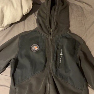 Mörkgrå hoodiejacka från Seal Arctic sail racing - Mörkgrå hoodiejacka med dragkedja och bröstficka från Seal Arctic. Jackan har en patch på vänster bröst och kontrasterande paneler över axlar och bröst. Perfekt för kyliga dagar och har en sportig look.