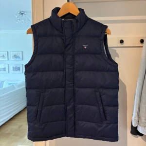 Gant dunväst - Snygg mörkblå dunväst från Gant med två fickor framtill i storlek 158/164 cm (13-14 år). Insidan är fodrad med vitt och blått randigt tyg. Perfekt för lager-på-lager-stil.