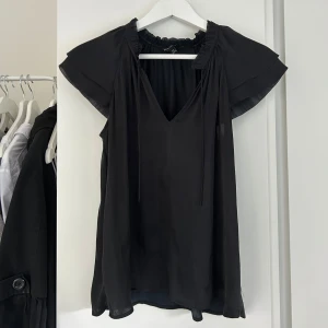 Svart blus med volangdetalj - Svart blus med korta, luftiga ärmar och v-ringning. Volangdetalj vid halsen och knytband. Tunn och flowy modell, perfekt för en stilren look.