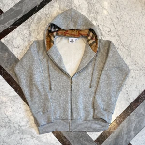 Burberry hoodie med dragkedja  - Säljer en grå burberry zip up med mönster i huvan. Den är helt ny men jag använde den ett fåtal gånger på semester. Säljer för att jag är sugen på nytt.