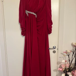 Röd långklänning med pärldetalj - Elegant röd långklänning med långa ärmar och vacker pärldetalj i midjan. Klänningen har draperad effekt framtill och ett flytande fall. Perfekt för speciella tillfällen när du vill sticka ut.