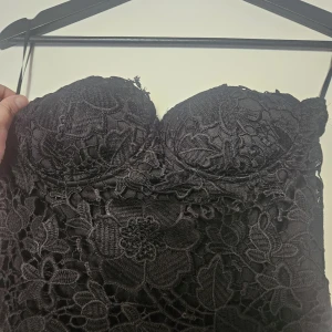 Svart långklänning med spets från Club L - Elegant svart långklänning från Club L med spetsdetaljer upptill och vadderad byst. Axelbandslös design och flowy kjol i tunt material. Perfekt för speciella tillfällen.