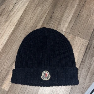 Svart stickad mössa från Moncler - Svart ribbstickad mössa från Moncler med uppvikt kant och klassisk Moncler-logga framtill. Tillverkad i mjukt material som håller värmen under kyliga dagar. Perfekt accessoar för en stilren look.