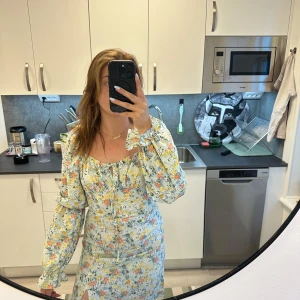 Blommig långklänning med öppen rygg och volang - Säljer en ljusblå långklänning med blommigt mönster i gult, orange och grönt. Klänningen har långa puffärmar, volangdetaljer vid halsringningen, slits framtill och dragkedja i ryggen. Perfekt för sommarens tillfällen! Den är tyvärr för liten på mig. Aldrig använd