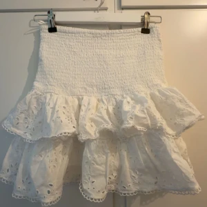 Vit volangkjol med broderade detaljer - Supersöt vit kortkjol med volanger och broderade hålmönster. Kjolen har smock i midjan för en bekväm passform och dubbla volanglager som ger extra volym. Perfekt för sommarens alla tillfällen! Men har en liten gul fläck på ❣️❣️