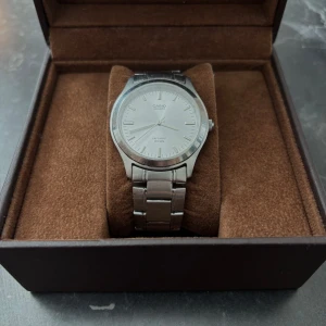 Silverfärgad klocka från Casio - Stilren analog klocka från Casio i helsilver med metallarmband och rund urtavla. Urtavlan är ljus med tydliga markeringar och smala visare. Passar dig som gillar klassisk och enkel design och helt nytt batteri. (box ingår ej)