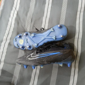 Svarta Nike fotbollsskor med blå detaljer - Säljer ett par svarta Nike fotbollsskor med blå och vita detaljer. Skorna har snörning och en platt sula med dobbar för bra grepp på planen. Perfekta för dig som vill ha stilrena och sportiga skor till matchen. Nike phantoms GX med griprint för bättre känsla. Lätta och sköna för intensivt spel. Storlek 40.5