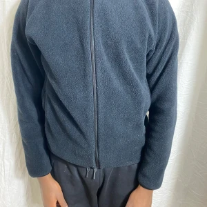 Svart fleecejacka med dragkedja - Mjuk och enkel svart fleecejacka med hel dragkedja framtill. Jackan har lång ärm och hög krage, perfekt för kyliga dagar. Passformen är normal och materialet är fleece som håller dig varm.