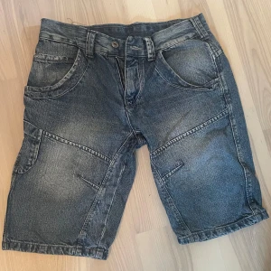 Blå jeansshorts från Across - Säljer ett par blå jeansshorts från Across med coola sömdetaljer och stora bakfickor med knappar. Klassisk femficksmodell och bälteshällor. Perfekta för sommaren och avslappnad stil.