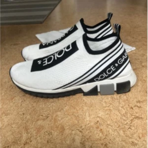 Dolce & Gabbana vita sneakers med svart logga - Säljer ett par vita Dolce & Gabbana sneakers med svart logotyp på ovansidan och sidan. Skorna har elastisk ovandel i textil, svart-vit randig hälflik och kraftig, mönstrad sula. Snygg och sportig design med slip-on modell.