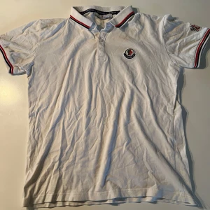 Vit piké t-shirt från Moncler - Vit piké t-shirt från Moncler med broderad logga på bröstet. Kragen och ärmsluten har röd, vit och blå randig detalj. Klassisk passform och kortärmad modell, perfekt för en stilren look. Den översta knappen är dessvärre sönder men går enkelt att sy på egen.