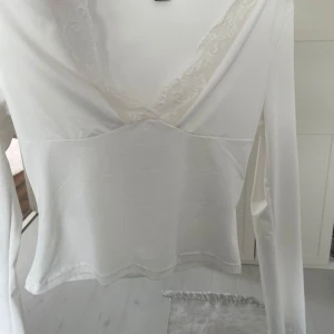 Vit omlottopp med spetsdetaljer - Säljer en vit omlottopp med långa ärmar och vacker spets längs den djupa v-ringningen. Toppen har en feminin och elegant look, perfekt att styla till olika outfits.