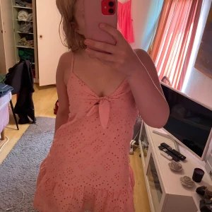 Rosa kortklänning med volang och knyt - Söt rosa kortklänning med broderade hålmönster, smala axelband och volang nertill. Klänningen har en knytning framtill vid bysten och är perfekt för varma dagar.