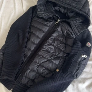 Moncler jacka - Säljer en svart pufferjacka från Moncler med stickade ärmar och huva. Jackan har dragkedja framtill och en ficka med dragkedja på ärmen. Perfekt för kyliga dagar och riktigt snygg streetstil.