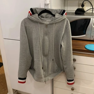  Moncler - Säljer en stilren grå hoodie från Moncler med dragkedja och randiga detaljer i rött, vitt och blått på ärmslut och huva. Perfekt för en avslappnad look. Riktigt snygg och inga fläckar eller liknande. Hör av er vid funderingar. Är storlek XXL men passar storlek L perfekt