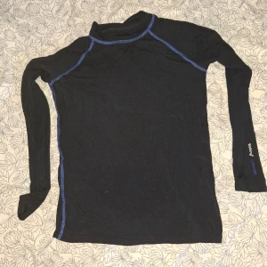 Svart långärmad träningströja från Walking - Svart långärmad träningströja från Walking med blå sömmar. Perfekt för utomhusaktiviteter och träning. Tillverkad i ett syntetmaterial som andas och transporterar bort fukt. Enkel och sportig design.