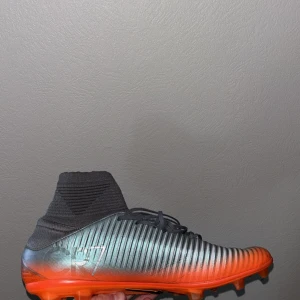 Nike Mercurial fotbollsskor i silver och orange - Säljer ett par Nike Mercurial fotbollsskor med högt skaft i silver och orange. Skorna har snörning, svart Nike-logga och platt sula med dobbar för gräsplan. Perfekta för dig som vill sticka ut på planen! Passar 42 också