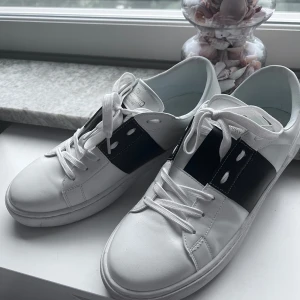 Valentino Garavani vita sneakers - Snygga vita sneakers från Valentino Garavani med bred svart rem över vristen och coola nitar på hälen. Skorna har rund tå, snörning och är tillverkade i skinn för en lyxig känsla. Perfekta för dig som gillar stilrena och exklusiva sneakers.