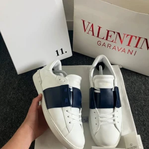 Valentino Garavani Open sneakers vit/blå - Snygga Valentino Garavani Open sneakers i vitt skinn med bred marinblå läderdetalj över vristen. Klassisk rund tå, platt sula och vita snören. Ikonisk och stilren design som sticker ut med den kontrasterande blåa remmen.