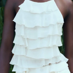 Vit volangig ärmlös blus - Supersöt vit blus med flera lager volanger och ärmlös design. Toppen har en halterneck-skärning och är tillverkad i ett lätt, luftigt tyg som ger en romantisk och trendig känsla. Perfekt för varma dagar när du vill sticka ut lite extra.