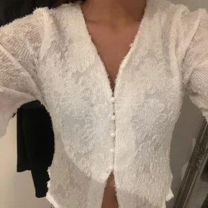 Vit spetsblus med pärlknappar - Supersöt vit blus i spets med v-ringning och långa ärmar. Blusen har fina pärlknappar framtill och lätt utsvängda ärmslut. Materialet är genombrutet och ger en romantisk vibe, perfekt att styla med jeans eller kjol.