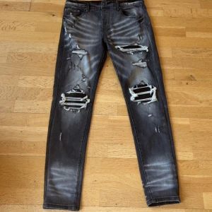 Svarta ripped jeans från Amiri - Säljer ett par svarta skinny jeans från Amiri med slitna detaljer och stora hål på benen. Jeansen har en tvättad look med ljusa partier och klassisk femficksmodell. Perfekta för dig som gillar streetstyle och edgy vibe.