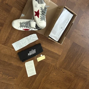 Limited edition Golden Goose  - Golden Goose sneakers i vitt skinn med grå snörning, röd stjärna på sidan och svart detalj på hälen. Klassisk rund tå och platt sula. Snygg och ikonisk modell som sticker ut med sin röda stjärndetalj. 